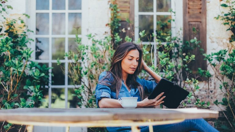 Eine Person sitzt im Freien vor einem Haus an einem Tisch und liest auf einem eReader.