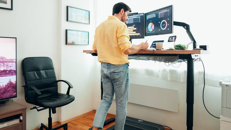 Eine Person geht auf einem Walking-Pad unter einem h&ouml;henverstellbaren Schreibtisch, auf dem ein professionelles Office Set-Up steht.