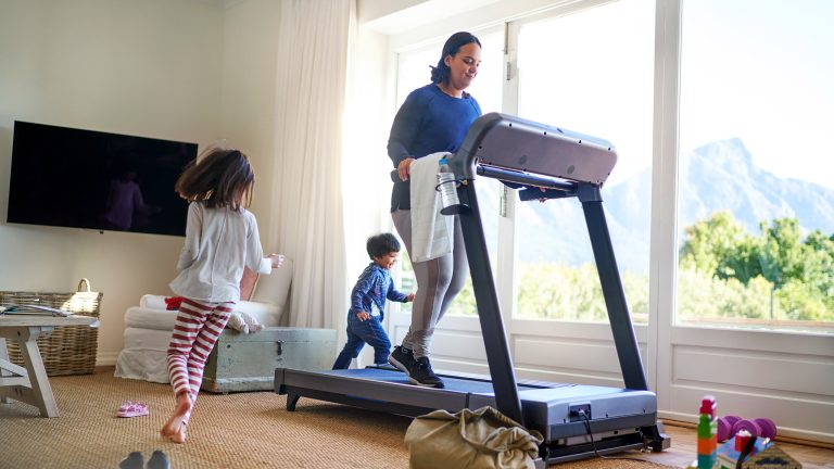 Eine Person trainiert im Wohnzimmer auf einem Laufband. Drumherum spielen zwei Kinder.