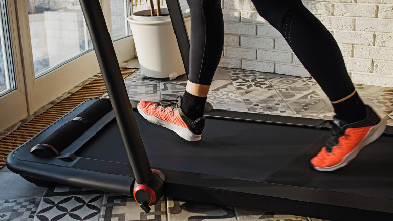 Es sind die Beine einer Person zu sehen, die in Sportkleidung und -schuhen stecken und auf einem Walking-Pad vor einer Terrassent&uuml;r gehen.