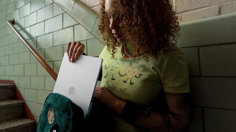 Eine Person steht in einem relativ dunklen Schulflur an einer Treppe und packt das MacBook Air in ihren Rucksack.