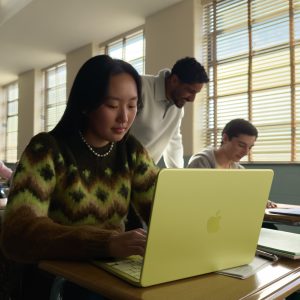 Eine Person sitzt vor einem MacBook Neo in der Farbe Zitrus in eiem Klassenzimmer.