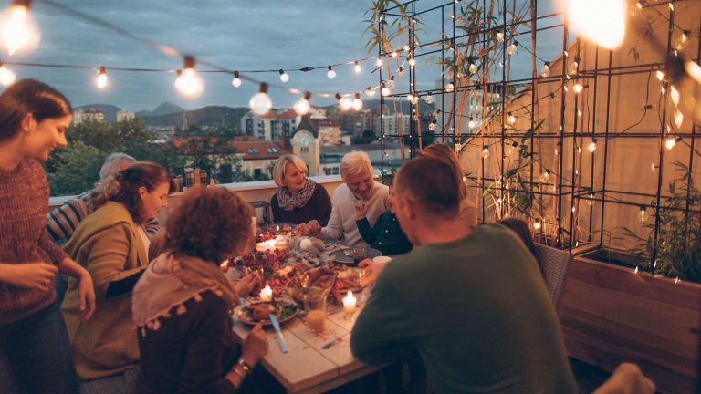 Balkon gestalten und bepflanzen, So wird er zu deinem Lieblingsplatz Eine Gruppe von Menschen sitzt abends um einen festlich gedeckten Tisch auf einem Dachbalkon. Ringsum hängen warmweiße Lichterketten, an der Seite ein begrüntes Rankgitter. Im Hintergrund der abendliche Stadthorizont.