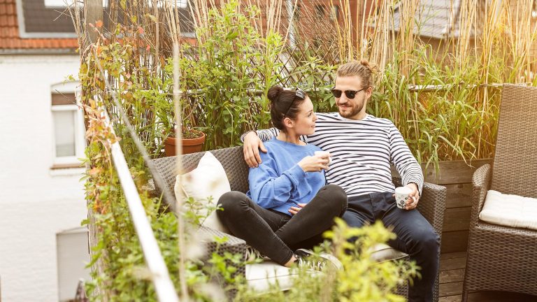 Zwei Personen sitzen eng beieinander auf einem Polyrattan-Sofa auf einem Balkon. Im Hintergrund ein nat&uuml;rlicher Sichtschutz aus hohen Gr&auml;sern und Pflanzk&uuml;beln, dahinter H&auml;user.