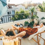 Zwei Personen liegen entspannt auf Liegest&uuml;hlen auf einem begr&uuml;nten Stadtbalkon. Rundherum stehen Topfpflanzen, an der Balkonbr&uuml;stung h&auml;ngen warmwei&szlig;e Gl&uuml;hbirnen. Im Hintergrund H&auml;user und blauer Himmel.