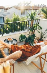 Balkon gestalten und bepflanzen, So wird er zu deinem Lieblingsplatz Zwei Personen liegen entspannt auf Liegestühlen auf einem begrünten Stadtbalkon. Rundherum stehen Topfpflanzen, an der Balkonbrüstung hängen warmweiße Glühbirnen. Im Hintergrund Häuser und blauer Himmel.
