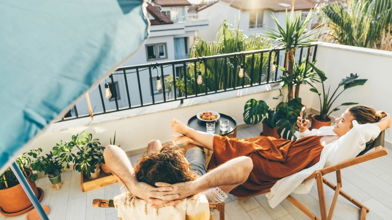 Zwei Personen liegen entspannt auf Liegest&uuml;hlen auf einem begr&uuml;nten Stadtbalkon. Rundherum stehen Topfpflanzen, an der Balkonbr&uuml;stung h&auml;ngen warmwei&szlig;e Gl&uuml;hbirnen. Im Hintergrund H&auml;user und blauer Himmel.
