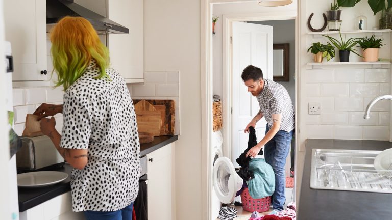 Eine Person l&auml;dt im Hintergrund eine Waschmaschine mit W&auml;sche voll. Im Vordergrund steht eine weitere Person in einer K&uuml;che und macht Toast.