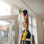 Eine Person steht auf einer Leiter und reinigt den Rahmen eines Fensters mit einem Tuch. Dabei tr&auml;gt sie gelbe Gummihandschuhe.