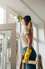 Fenster streifenfrei putzen, So klappt’s endlich Eine Person steht auf einer Leiter und reinigt den Rahmen eines Fensters mit einem Tuch. Dabei trägt sie gelbe Gummihandschuhe.