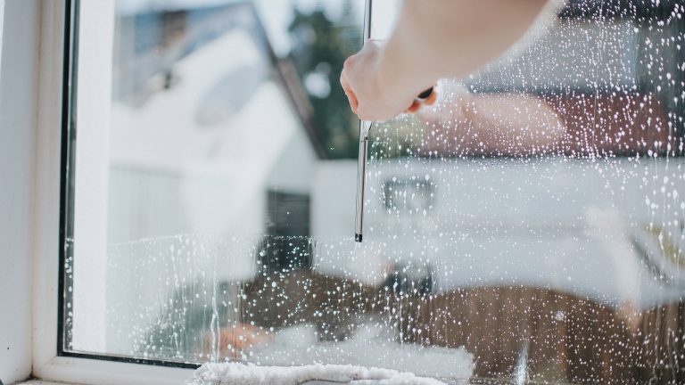 Fenster streifenfrei putzen, So klappt’s endlich Detailaufnahme eines eingeschäumten Fensters. Es ist der Arm einer Person zu sehen, die den reinigenden Schaum mithilfe eines Gummiabziehers entfernt.