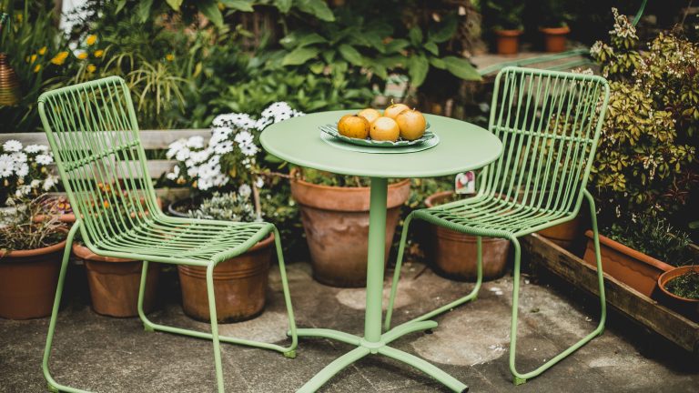 Gartenmöbel reinigen, schonend und leicht mit cleveren Gadgets Ein Set aus zwei Metallstühlen und einem Tisch steht in der Ecke einer Terrasse mit vielen Blumentöpfen und Pflanzen im Hintergrund.