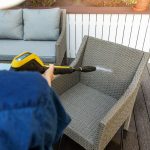 Blick &uuml;ber die Schulter einer Person, die auf einer Terrasse einen Gartenstuhl aus Rattan mit einem Hochdruckreiniger bespr&uuml;ht.