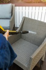 Gartenmöbel reinigen, schonend und leicht mit cleveren Gadgets Blick über die Schulter einer Person, die auf einer Terrasse einen Gartenstuhl aus Rattan mit einem Hochdruckreiniger besprüht.
