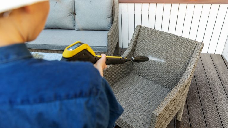 Blick &uuml;ber die Schulter einer Person, die auf einer Terrasse einen Gartenstuhl aus Rattan mit einem Hochdruckreiniger bespr&uuml;ht.