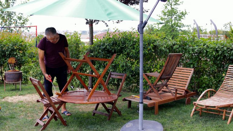 Eine Person steht in einem Garten unter einem Sonnenschirm und lackiert die Unterseite eines Holztisches, der umgekehrt auf zwei Holzst&uuml;hlen steht. Daneben sind weitere Holzm&ouml;bel.