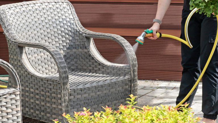 Eine Person spr&uuml;ht mit einer leichten Brause aus einem Gartenschlauch einen Gartenstuhl aus Rattangeflecht ab.
