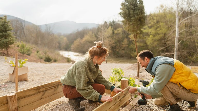 hochbeet-anlegen-4 Zwei Personen hocken nebeneinander und bauen gemeinsam einen Holzrahmen für ein Hochbeet zusammen. Eine Person hält ein Brett, die andere setzt einen Akkuschrauber an. Im Hintergrund eine Berglandschaft.