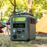 Im Vordergrund steht eine Powerstation. Sie steht im Gras. Im Hintergrund sind Zelte und ein Camping-Stuhl zu sehen.