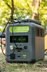 Im Vordergrund steht eine Powerstation. Sie steht im Gras. Im Hintergrund sind Zelte und ein Camping-Stuhl zu sehen.