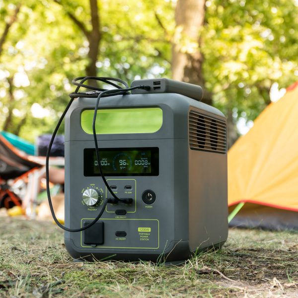 Im Vordergrund steht eine Powerstation. Sie steht im Gras. Im Hintergrund sind Zelte und ein Camping-Stuhl zu sehen.