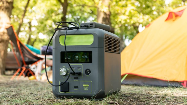 Im Vordergrund steht eine Powerstation. Sie steht im Gras. Im Hintergrund sind Zelte und ein Camping-Stuhl zu sehen.