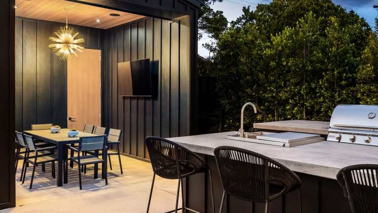 Eine modern eingerichtete Outdoor-K&uuml;che mit schwarzen Holzvert&auml;felungen und hellen Holzelementen, wie T&uuml;r und St&uuml;hle unter einer Pergola. Im Vordergrund ist eine steinerne Arbeitsplatte mit Sp&uuml;lbecken und Grill zu sehen.