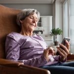 Eine &auml;ltere Person sitzt in einem Loungesessel und h&auml;lt dabei ein Smartphone in den H&auml;nden. Im Hintergrund ist ein Wohnzimmerschrank und eine Pflanze zu sehen.
