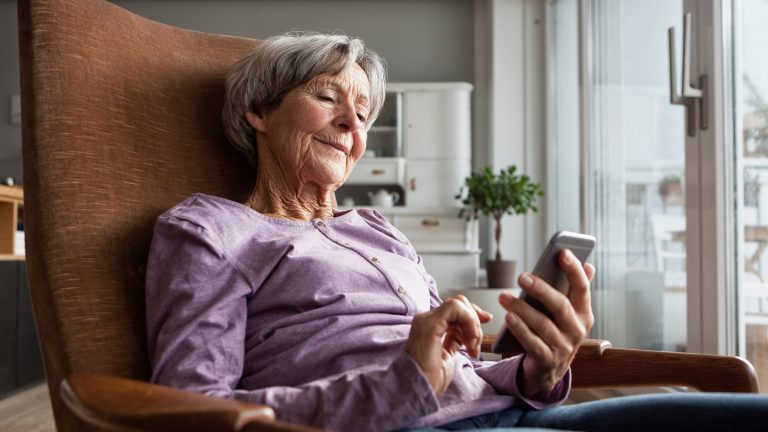 Eine &auml;ltere Person sitzt in einem Loungesessel und h&auml;lt dabei ein Smartphone in den H&auml;nden. Im Hintergrund ist ein Wohnzimmerschrank und eine Pflanze zu sehen.