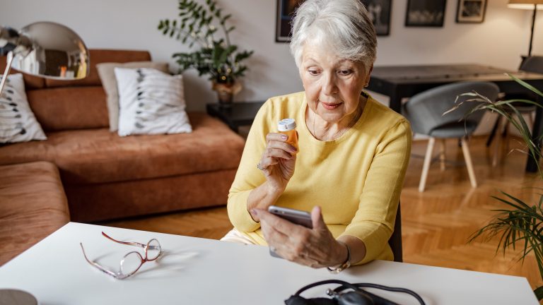 Eine &auml;ltere Person sitzt an einem Tisch im Wohnzimmer. In der einen Hand h&auml;lt sie eine Tablettendose, in der anderen ein Smartphone. Auf dem Tisch liegt eine Brille und ein Blutdruckmessger&auml;t.