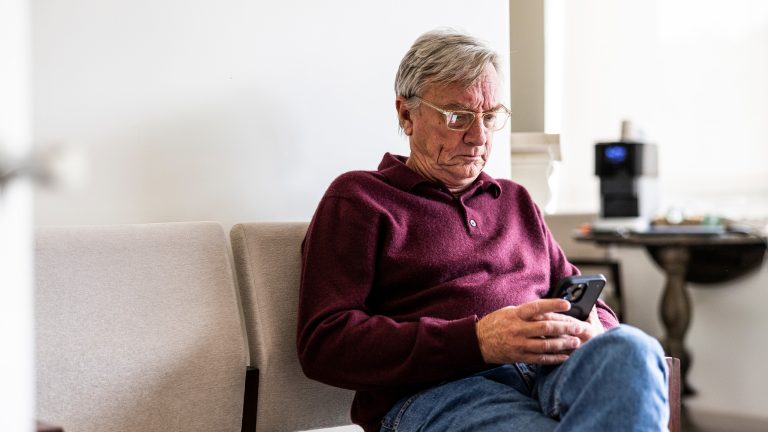 Eine &auml;ltere Person sitzt mit verschr&auml;nkten Beinen auf einem Stuhl und bedient ihr Smartphone. Im Hintergrn ist ein Tisch mit einer Kaffeemaschine zu erahnen.