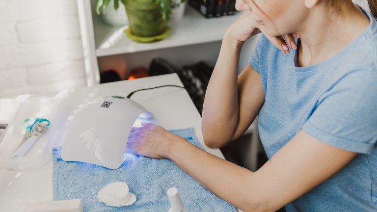 Eine Person sitzt an einem Kosmetiktisch. Darauf stehen Manik&uuml;re-Utensilien sowie eine Lampe, in die die Person ihre linke Hand zum Aush&auml;rten der N&auml;gel h&auml;lt.