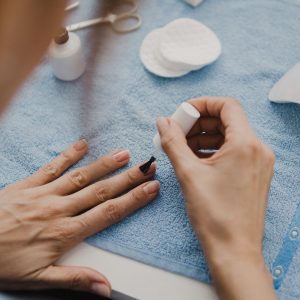 Vogelperspektive auf die linke Hand einer Person, die auf einem Kosmetiktisch ruht. Die rechte Hand der Person lackiert die Fingern&auml;gel mit Klarlack.