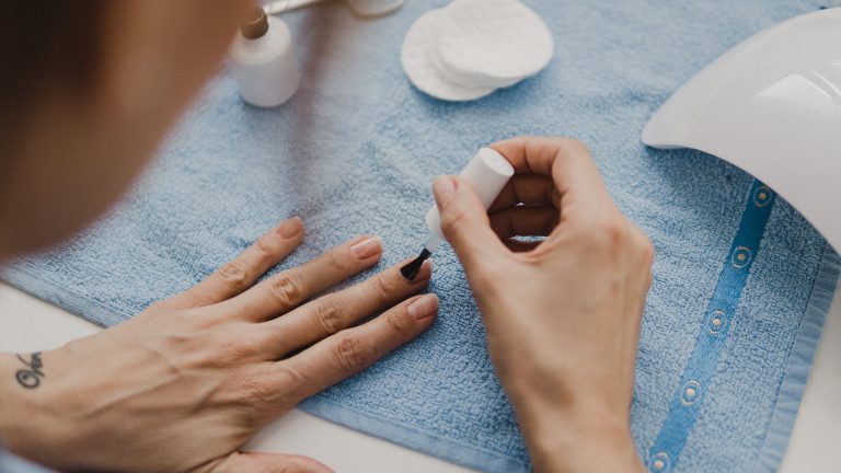 Vogelperspektive auf die linke Hand einer Person, die auf einem Kosmetiktisch ruht. Die rechte Hand der Person lackiert die Fingern&auml;gel mit Klarlack.