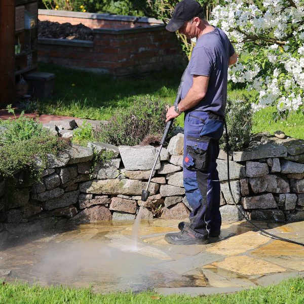 Eine Person in Arbeitskleidung reinigt eine Natursteintetrasse im Garten mit einem Hochdruckreiniger. Im Hintergrund bl&uuml;ht ein Apfelbaum.