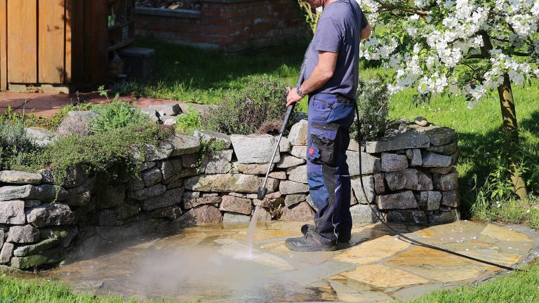 Eine Person in Arbeitskleidung reinigt eine Natursteintetrasse im Garten mit einem Hochdruckreiniger. Im Hintergrund bl&uuml;ht ein Apfelbaum.
