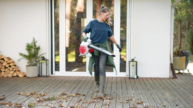 Eine Person mit Schutzbrille und Handschuhen bl&auml;st mit einem Laubbl&auml;ser Herbstlaub von einer verwitterten Holzterrasse vor einem Haus.