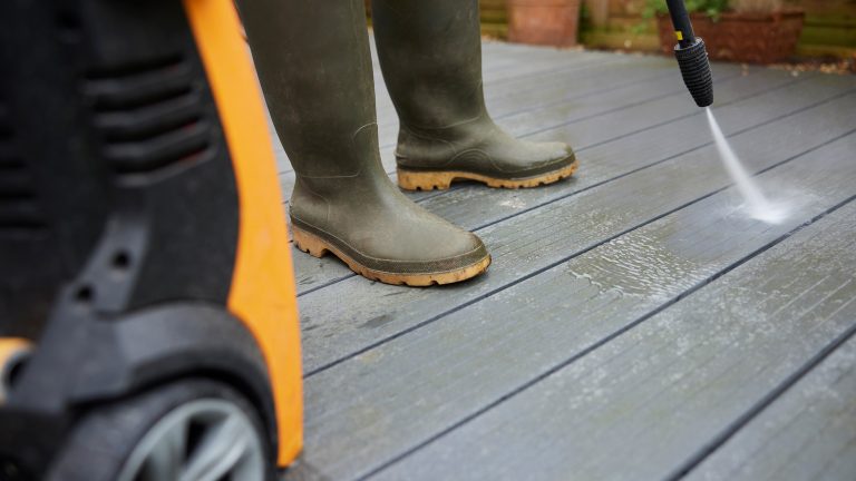 Nahaufnahme einer Person in Gummistiefeln, die mit dem Strahlrohr eines Hochdruckreinigers eine graue WPC-Terrasse reinigt. Im Vordergrund ist das Ger&auml;t mit orangefarbenem Geh&auml;use zu sehen.