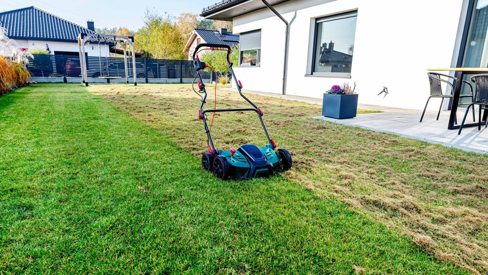 Ein elektrischer Vertikutierer steht auf einem gr&uuml;nen Rasen vor einem modernen Einfamilienhaus mit Terrasse. Im Hintergrund ist ein Garten mit herbstlicher Bepflanzung zu sehen.