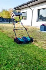 Rasen vertikutieren, So machst Du es richtig Ein elektrischer Vertikutierer steht auf einem grünen Rasen vor einem modernen Einfamilienhaus mit Terrasse. Im Hintergrund ist ein Garten mit herbstlicher Bepflanzung zu sehen.