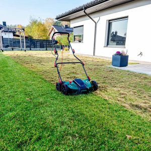 Ein elektrischer Vertikutierer steht auf einem gr&uuml;nen Rasen vor einem modernen Einfamilienhaus mit Terrasse. Im Hintergrund ist ein Garten mit herbstlicher Bepflanzung zu sehen.