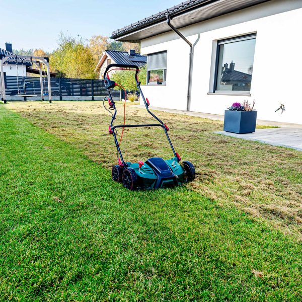 Ein elektrischer Vertikutierer steht auf einem gr&uuml;nen Rasen vor einem modernen Einfamilienhaus mit Terrasse. Im Hintergrund ist ein Garten mit herbstlicher Bepflanzung zu sehen.