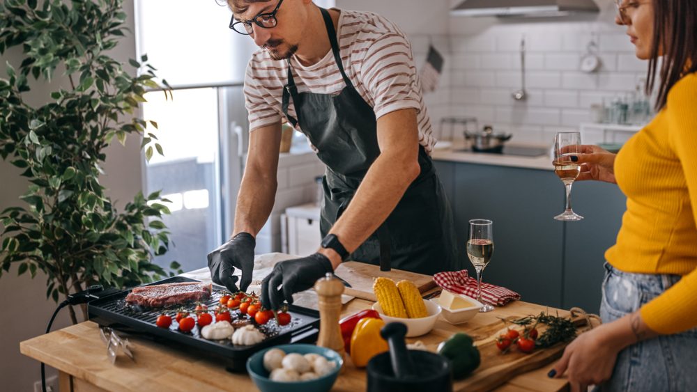 Eine Person in schwarzer Sch&uuml;rze und Handschuhen bereitet auf einem Elektrogrill Gem&uuml;se und Fleisch zu. Neben ihm steht eine Person mit einem Glas Wei&szlig;wein. Im Hintergrund eine helle, minimalistisch eingerichtete K&uuml;che mit Dunstabzugshaube.