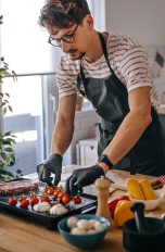 Eine Person in schwarzer Sch&uuml;rze und Handschuhen bereitet auf einem Elektrogrill Gem&uuml;se und Fleisch zu. Neben ihm steht eine Person mit einem Glas Wei&szlig;wein. Im Hintergrund eine helle, minimalistisch eingerichtete K&uuml;che mit Dunstabzugshaube.