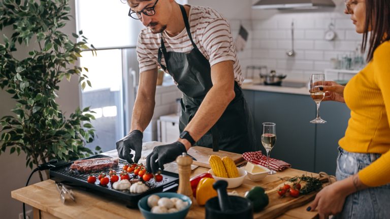 Eine Person in schwarzer Sch&uuml;rze und Handschuhen bereitet auf einem Elektrogrill Gem&uuml;se und Fleisch zu. Neben ihm steht eine Person mit einem Glas Wei&szlig;wein. Im Hintergrund eine helle, minimalistisch eingerichtete K&uuml;che mit Dunstabzugshaube.