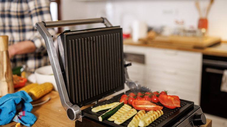 Ein Kontaktgrill mit buntem Gem&uuml;se steht auf einer Holzarbeitsplatte. Im Hintergrund bereitet eine Person im karierten Hemd weitere Zutaten vor.
