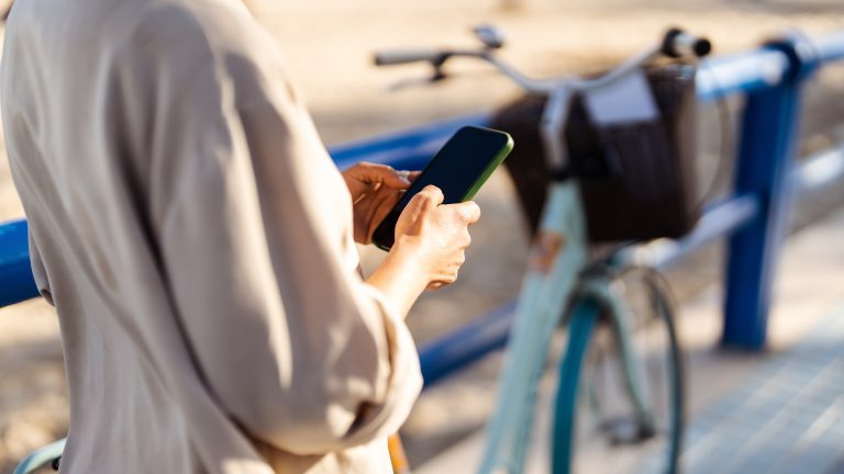 Halbnahe Aufnahme des Oberk&ouml;rpers einer Person, die ein Smartphone in der Hand h&auml;lt, im Hintergrund ist unscharf ein angekettetes Fahrrad mit Korb zu sehen.