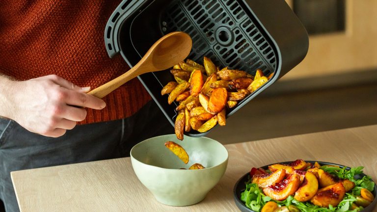Heißluftfritteuse richtig nutzen - Tipps für perfekte Ergebnisse Eine Person leert einen Garkorb mit gegarten Kartoffelspalten und Karotten in eine Schüssel, daneben ein Teller mit karamellisierten Pfirsichen auf Rucola.