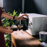 Eine Person h&auml;lt eine schwarze Kunststoff-Aufbewahrungsbox mit Reis und Beilagen vor eine wei&szlig;e Mikrowelle, um das Essen aufzuw&auml;rmen.