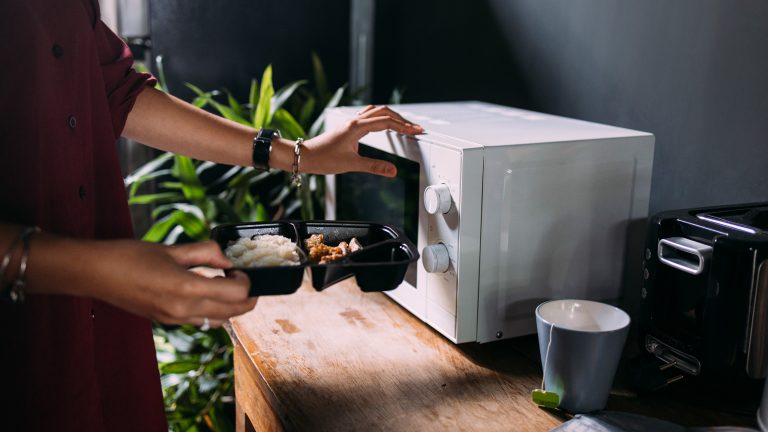 Eine Person h&auml;lt eine schwarze Kunststoff-Aufbewahrungsbox mit Reis und Beilagen vor eine wei&szlig;e Mikrowelle, um das Essen aufzuw&auml;rmen.